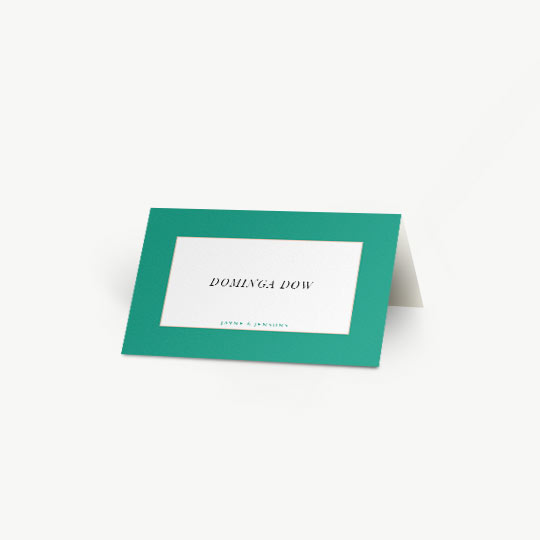 A bold, green place card personalised with the name of a wedding guest. The name of the bride and groom is printed at the bottom in blue. The folded place card sits on a white table.